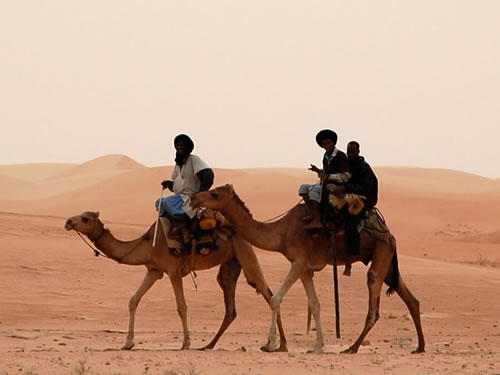 Three men carrying supplies on two camels across the desert.